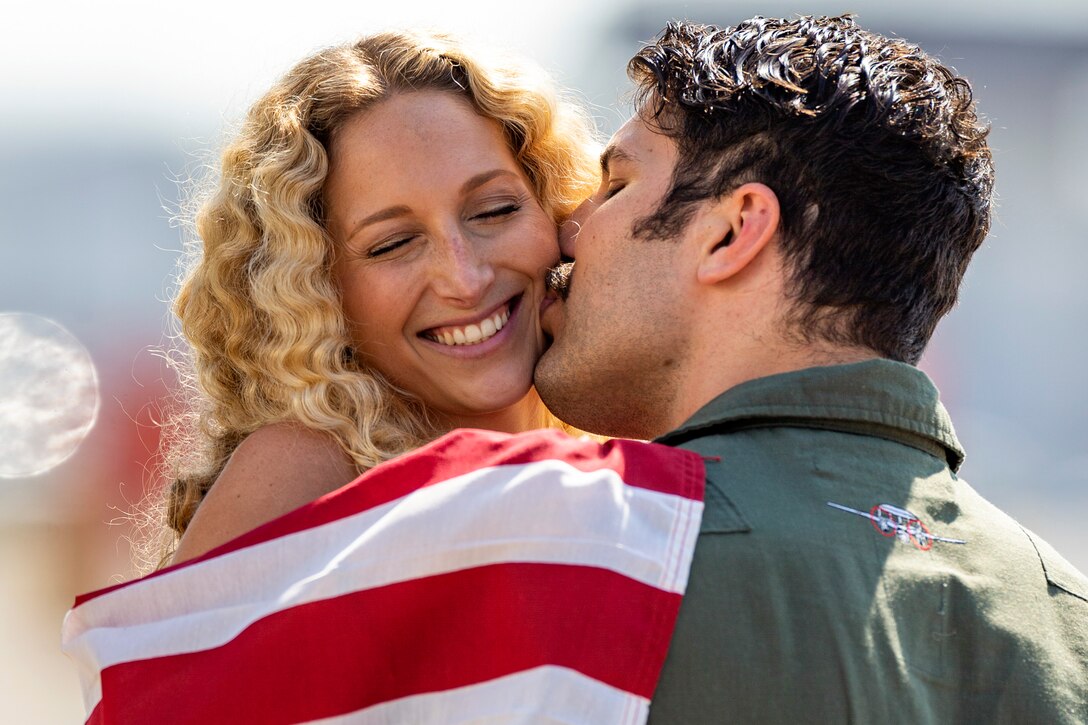 A sailor kisses his wife on the cheek while she holds an American flag.