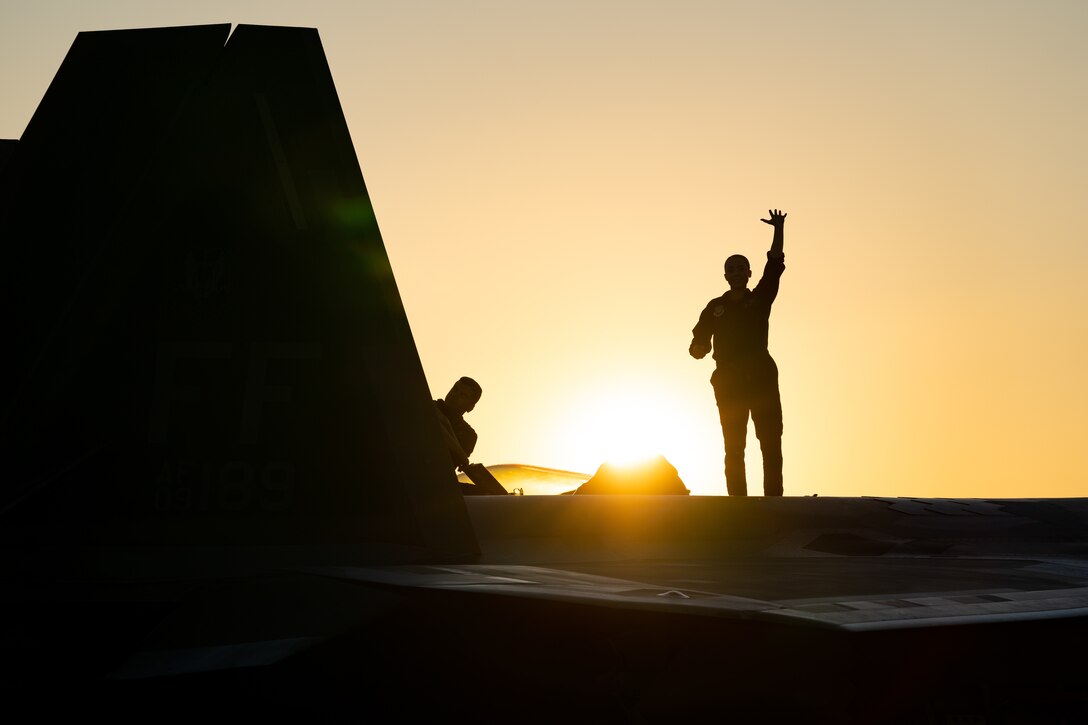 An airman waves while a fellow service member performs maintenance on an aircraft under a sunlit sky as seen in silhouette.