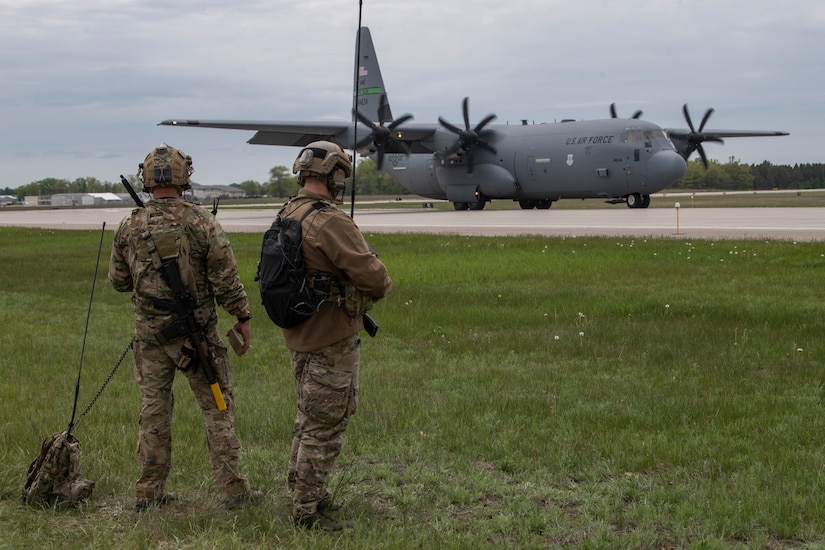 Two men in military uniforms and with communications equipment stand on a grassy area next to a runway where there is a large gray four-engine airplane.