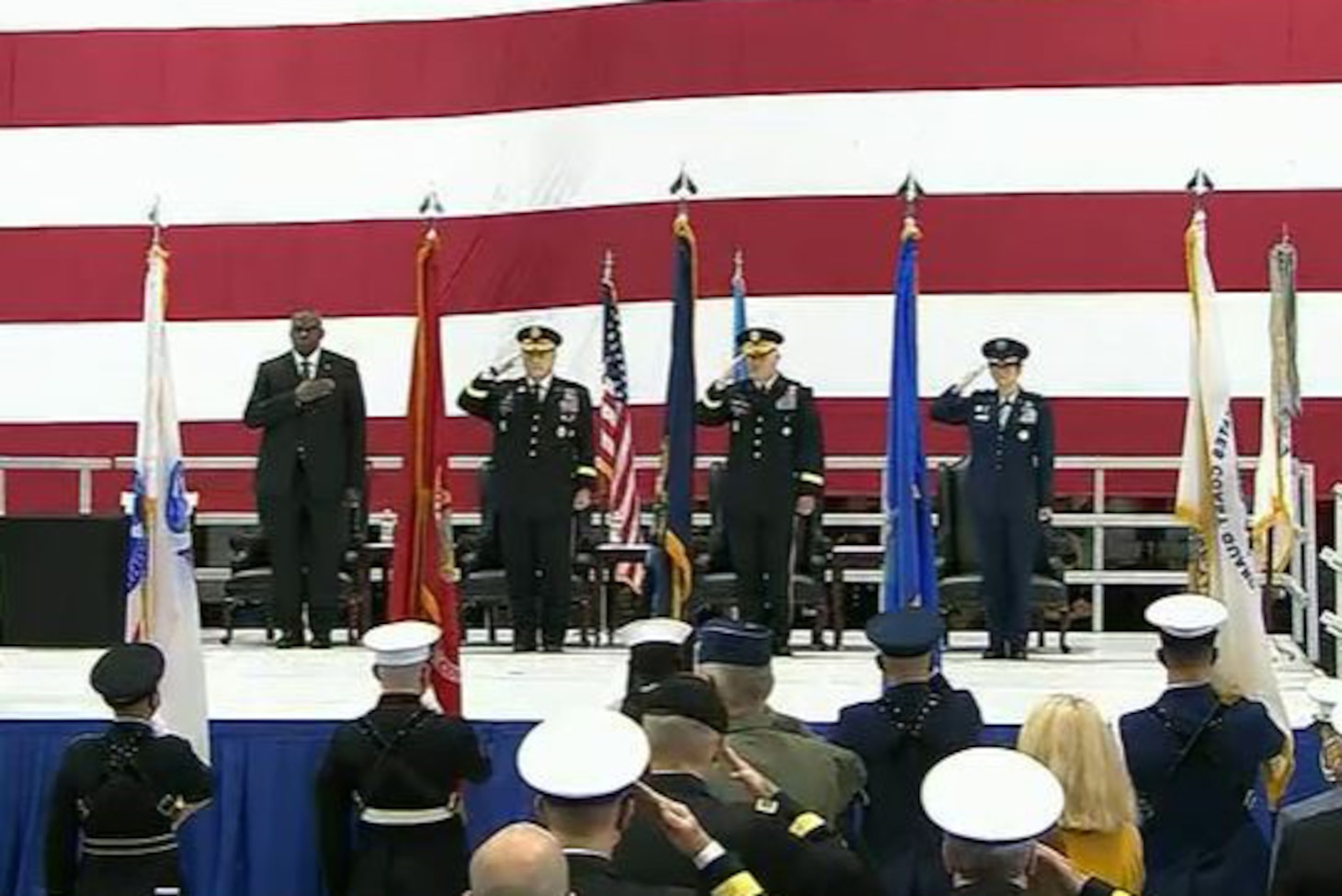 Defense Department leaders salute on a stage in front of a large American flag.