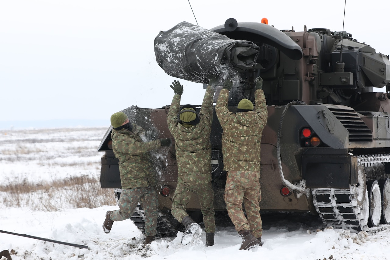 Three soldiers throw a large tarp in the back of a military vehicle moving through snow.