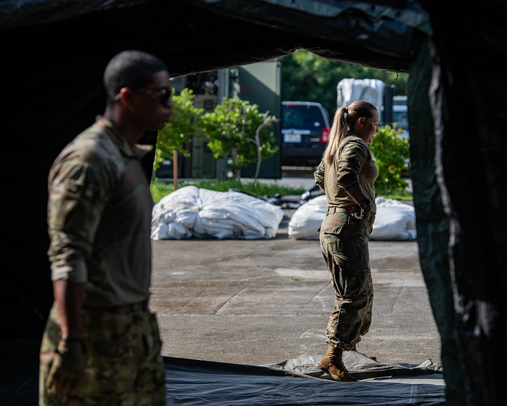 A man in the foreground walks through the light of the front of a tent while a woman in the background walks in the same direction