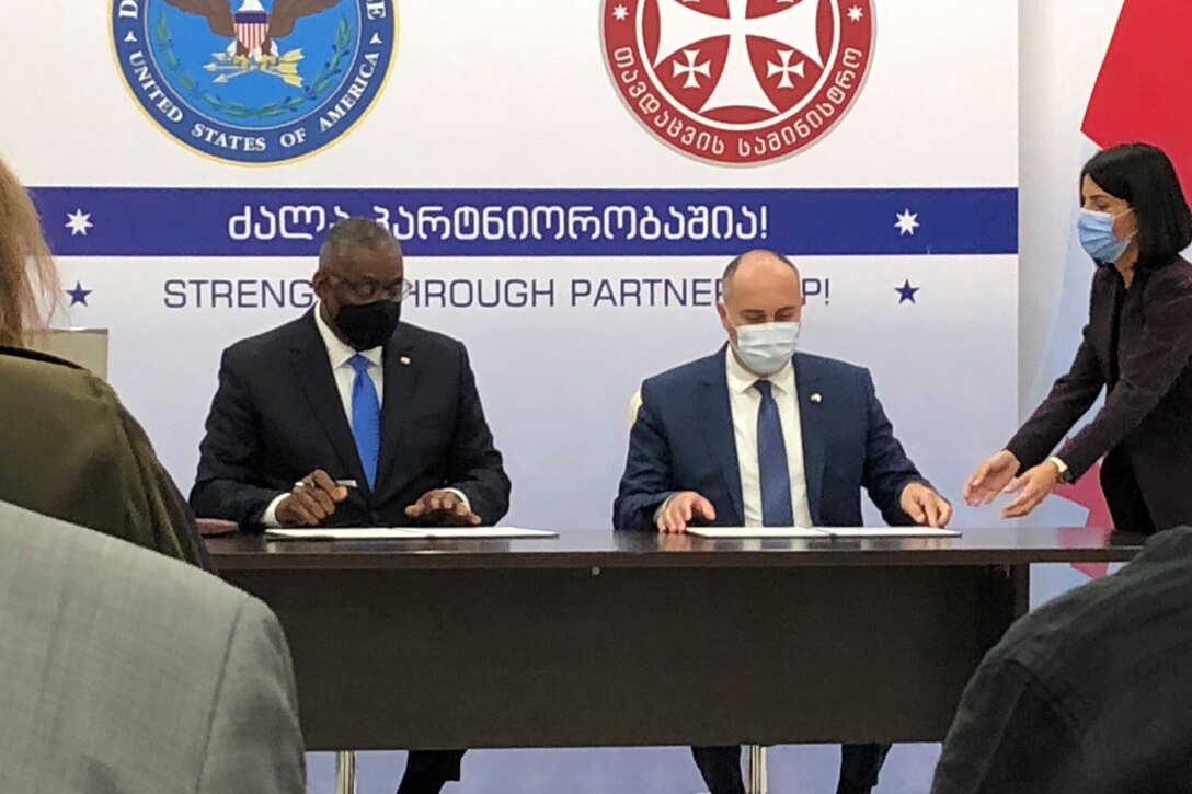 Two men sit at a table with papers in front of them.