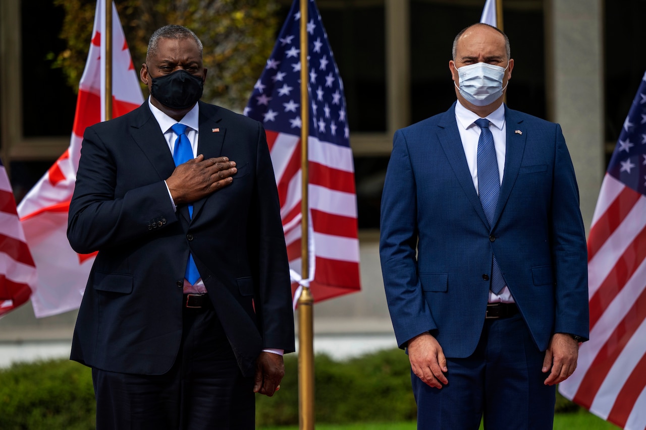 Two men stand in front of American flags, one holding his hand over his chest.