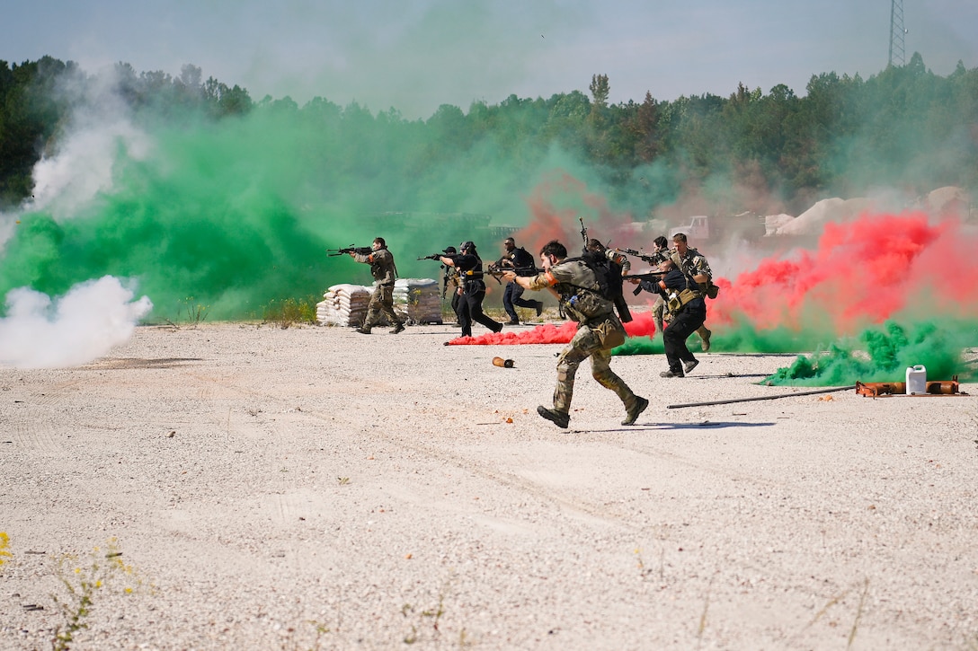 Soldiers aim weapons in a desert with clouds of red and green smoke in the background.