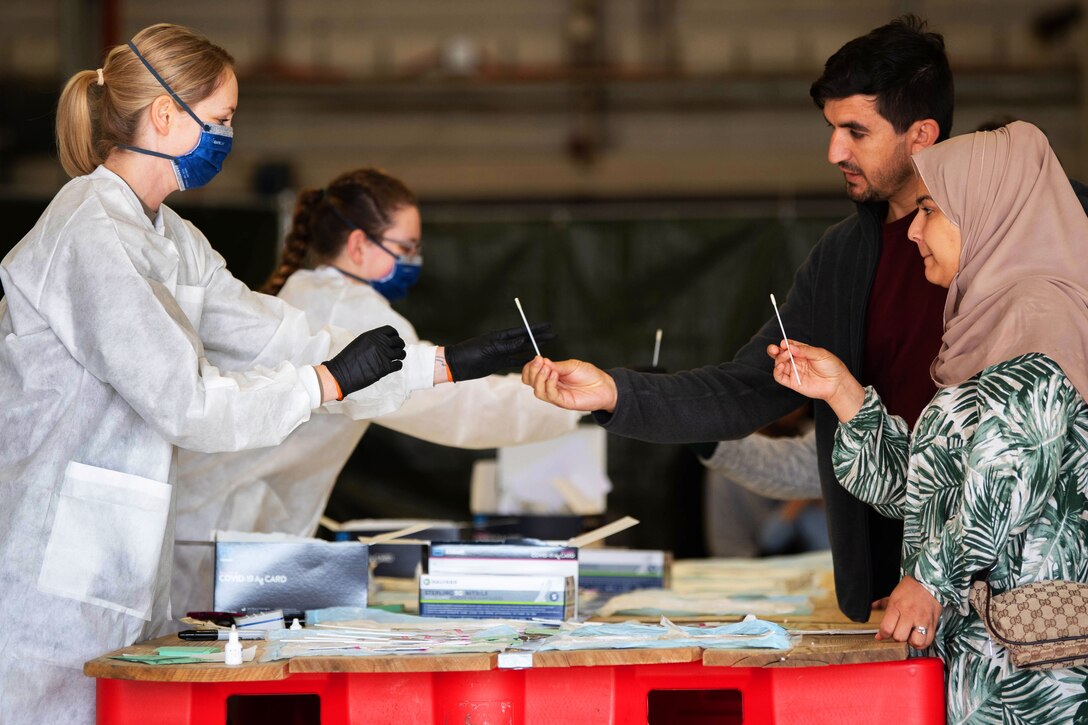 Two female airmen wearing face masks, gloves and medical gowns take nasal swabs from male and female Afghanistan evacuees.