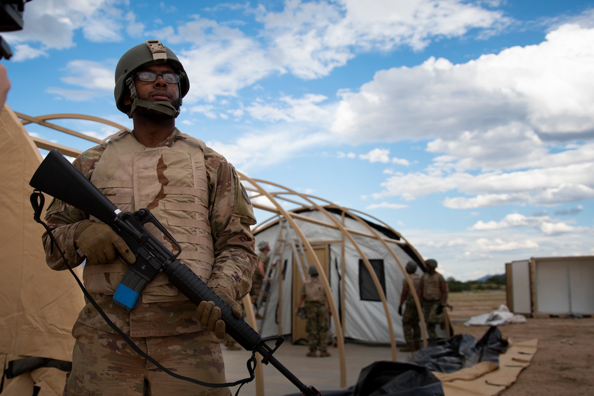 Man holds weapon while others build a tent behind him.