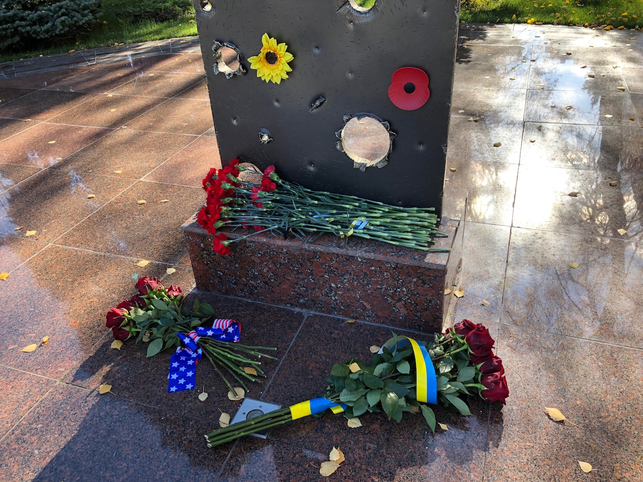 Bouquets of flowers are at the foot of a memorial pierced by armor rounds.