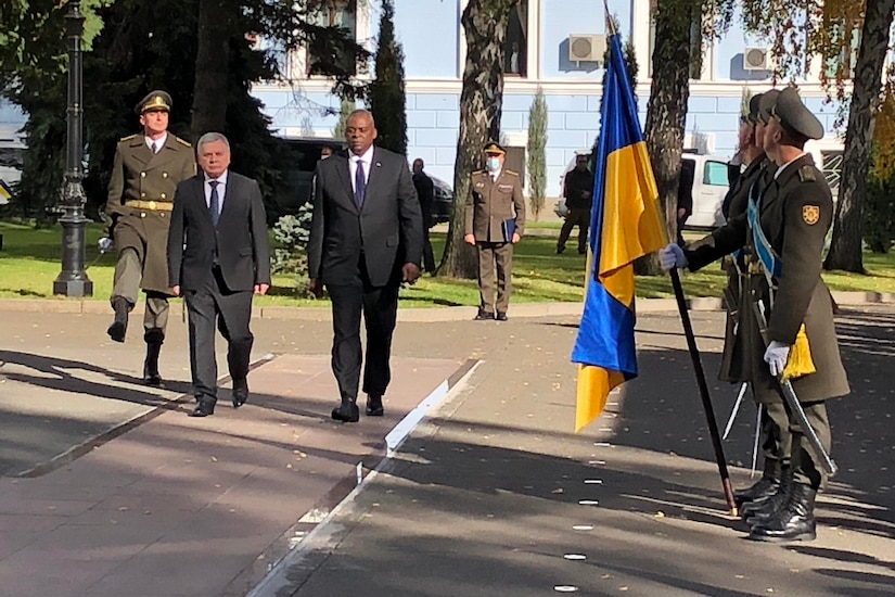 Two men dressed in business suits walk past soldiers holding flags.