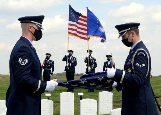 Two male Air Force service members fold an American flag at a cemetery while a four-person color guard stands in the background.