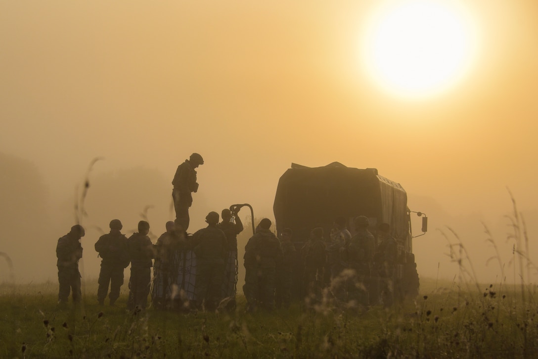 Soldiers attach cargo to a truck in a field under a sunlit sky.