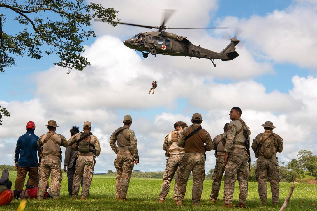 A soldier attached to an airborne helicopter hovers over a field as fellow soldiers watch.