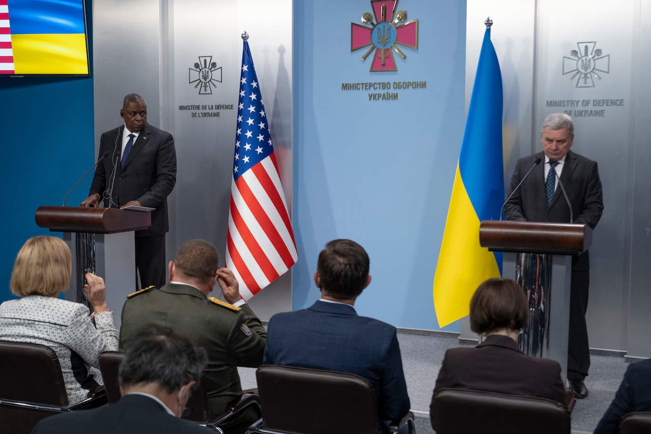 Two men stand on a stage in front of reporters.