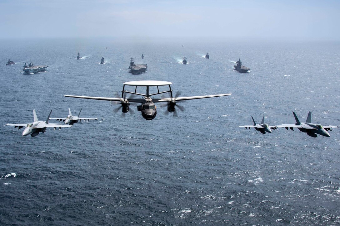 A large group of ships and aircraft move in formation across and above the water.