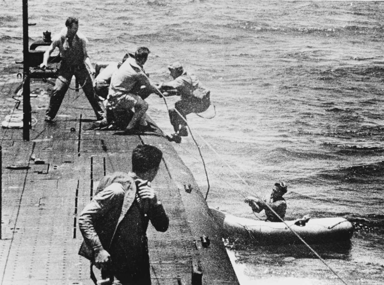 Several men on a surfaced submarine pull a man from a lifeboat. Another man sits in the boat.