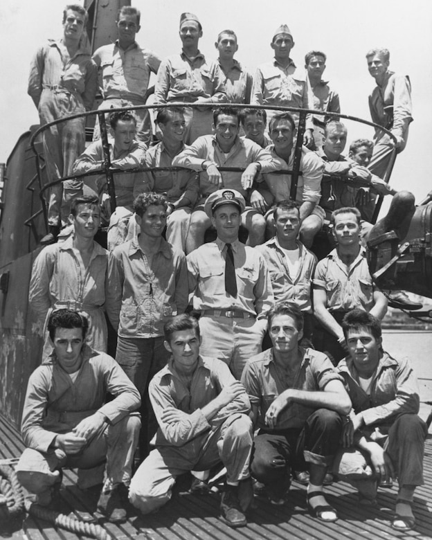 Several men pose for a photo in four rows on a ship's bridge.