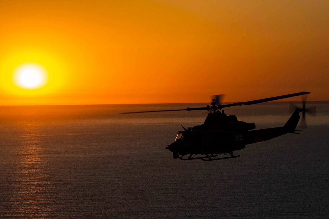 A Marine Corps helicopter flies as the sun shines in the background.