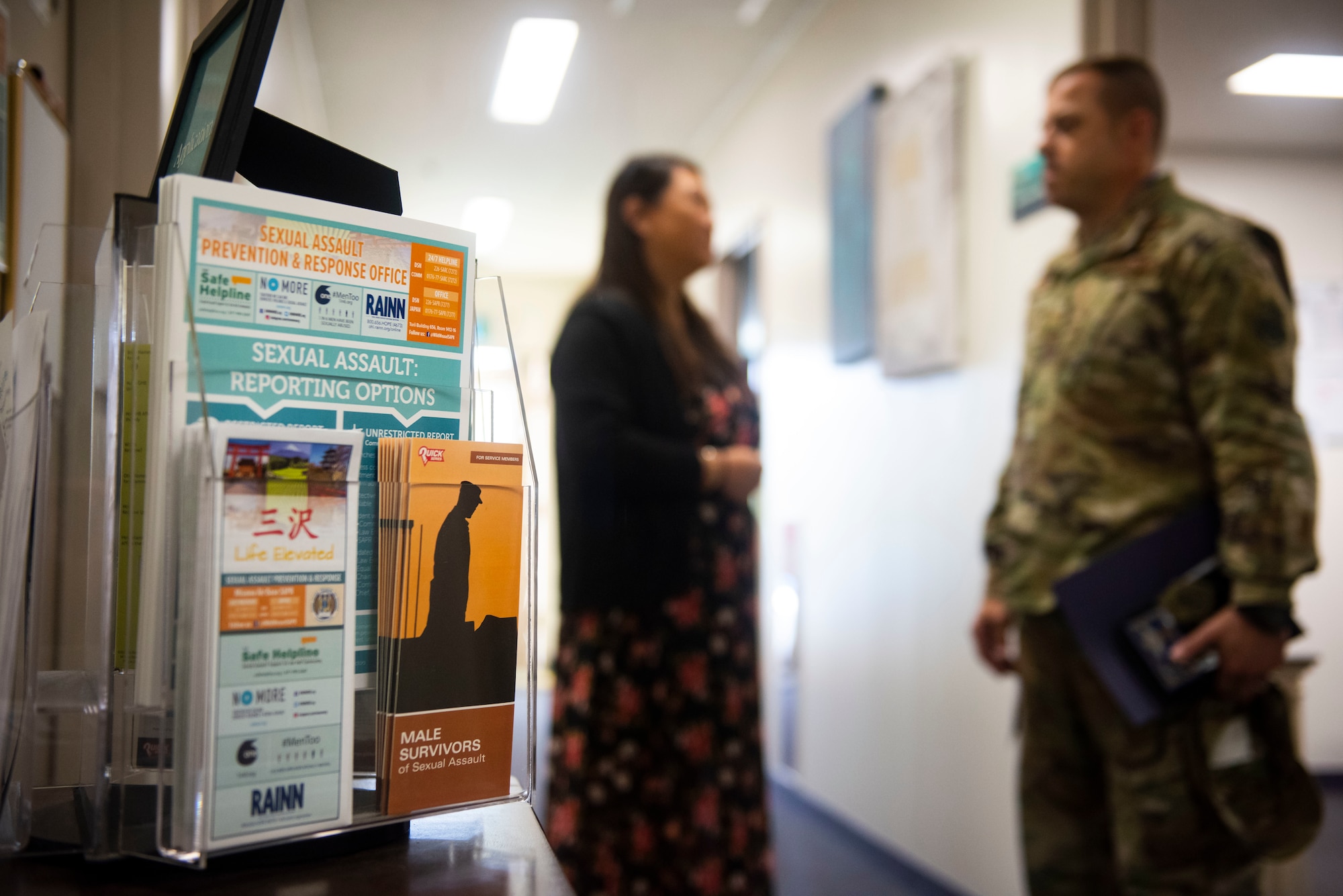 Pamphlets are on a table while man in military uniform stands and talks to female in the background.