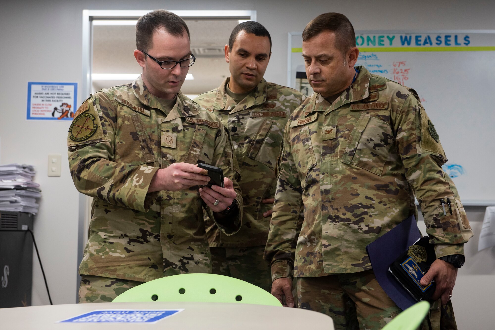 Three men in military uniform stand in an office looking at a phone.