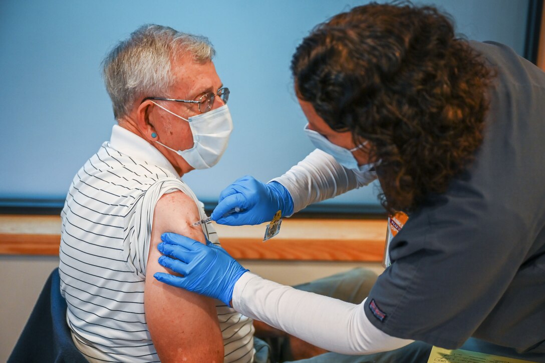 A woman wearing a face mask and gloves leans over while holding a syringe to give an elderly man, seated and wearing a face mask, a COVID-19 vaccine.