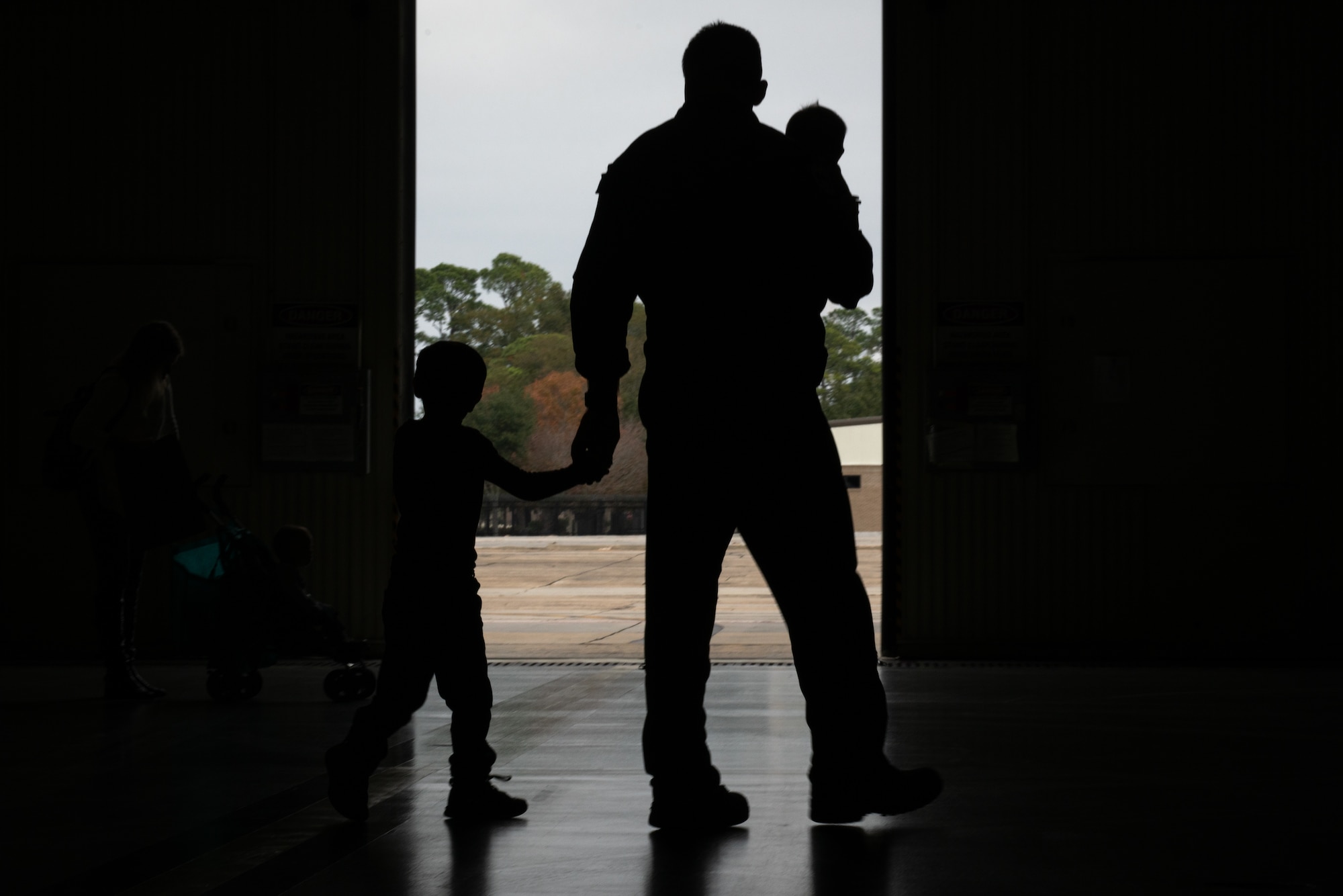 An Airman and holding their child, while holding the hand of another, are silhouetted in a hangar against the flightline background.