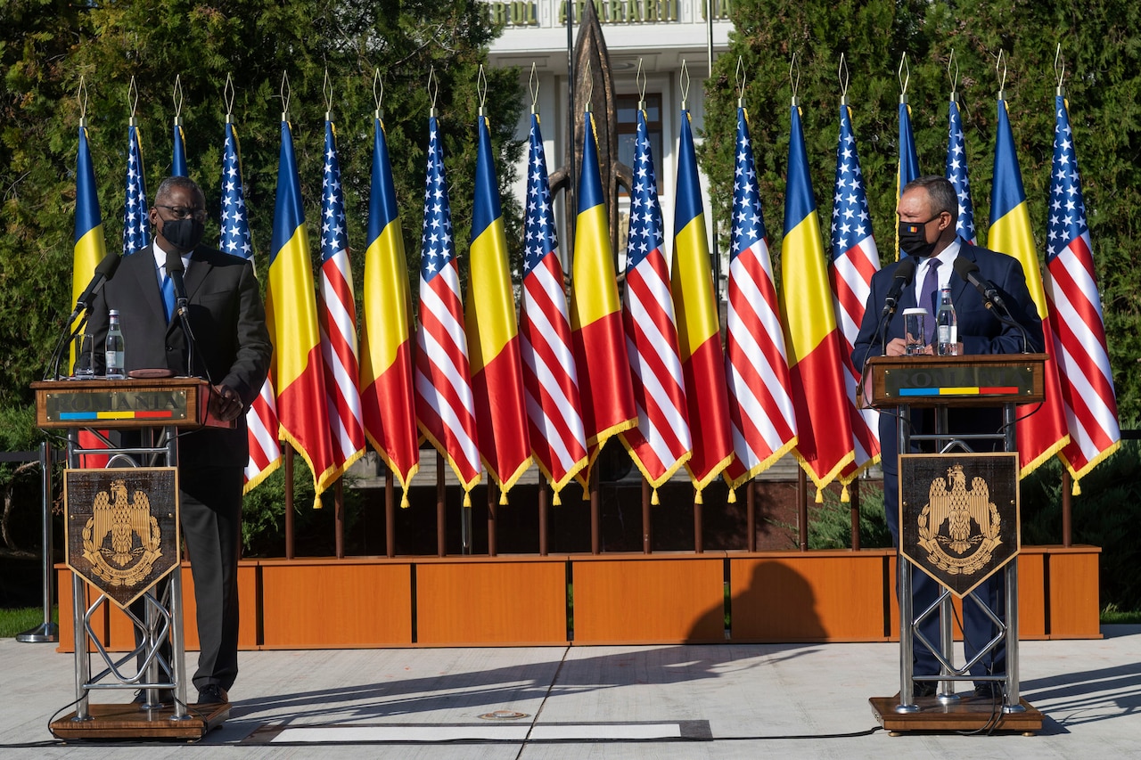 Two men stand in front of a row of flags and speak to reporters.
