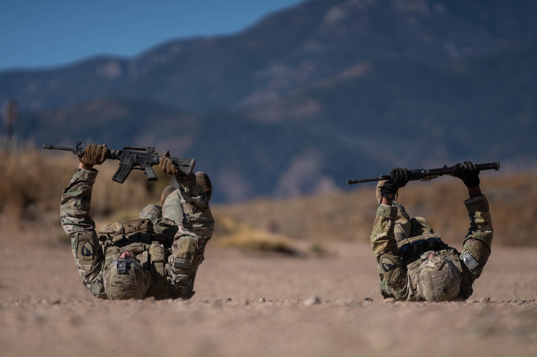 Two soldiers lie on the ground and hold their weapons above them.