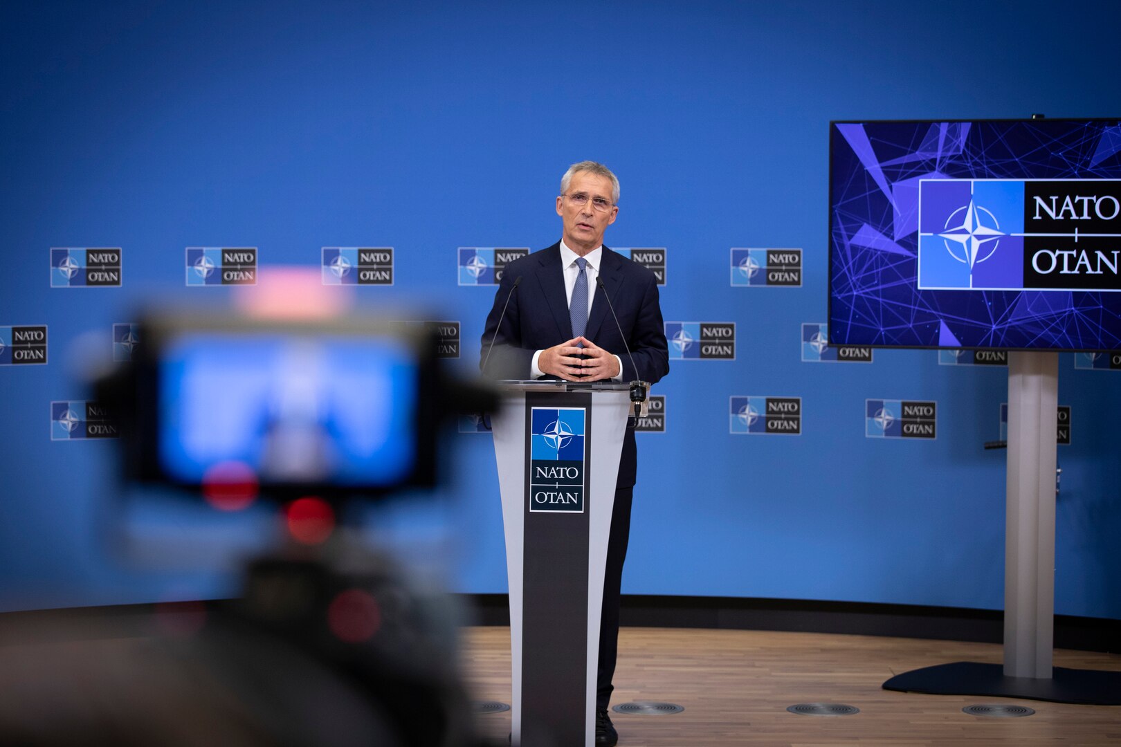A man stands at a podium. Behind him is a backdrop with repeated NATO symbols. In the foreground, the same man is visible on a camera monitor.