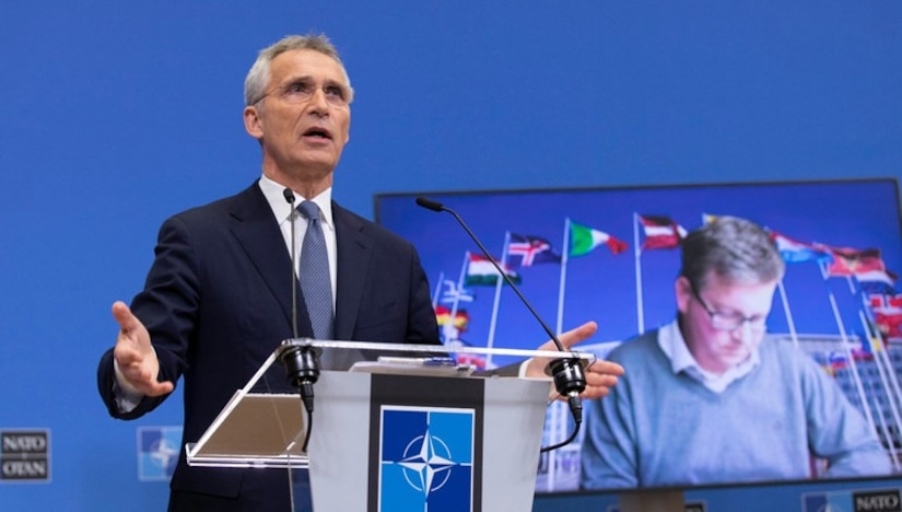 A man in a business suit stands at a lectern and speaks into a microphone. Behind him, a large screen shows another man looking downward.