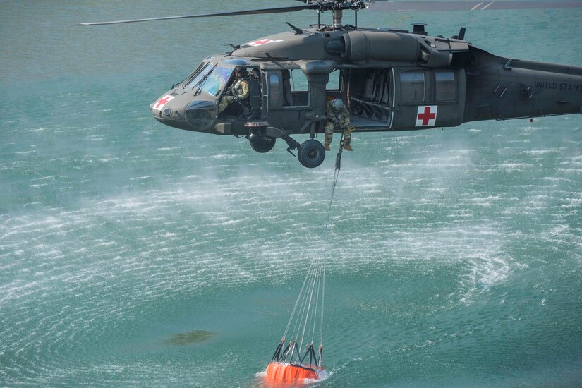 A helicopter and its crew lift a large bucket out of a body of water.