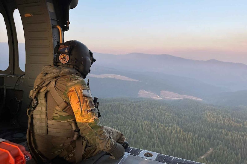 A person looks out the open door of an airborne helicopter at rolling hills and forest.