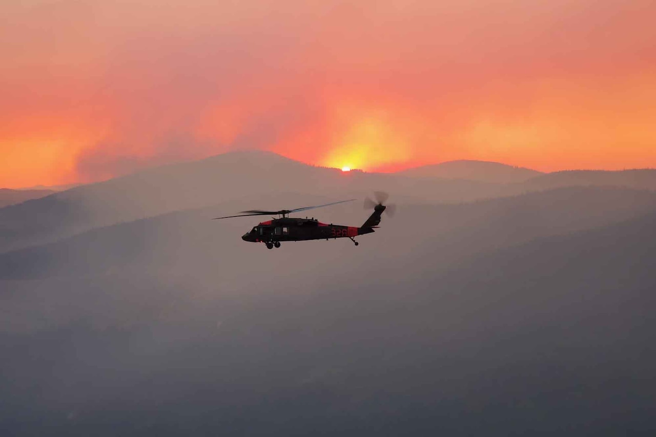 A helicopter flies over smoky mountains as the sun sets in the background.