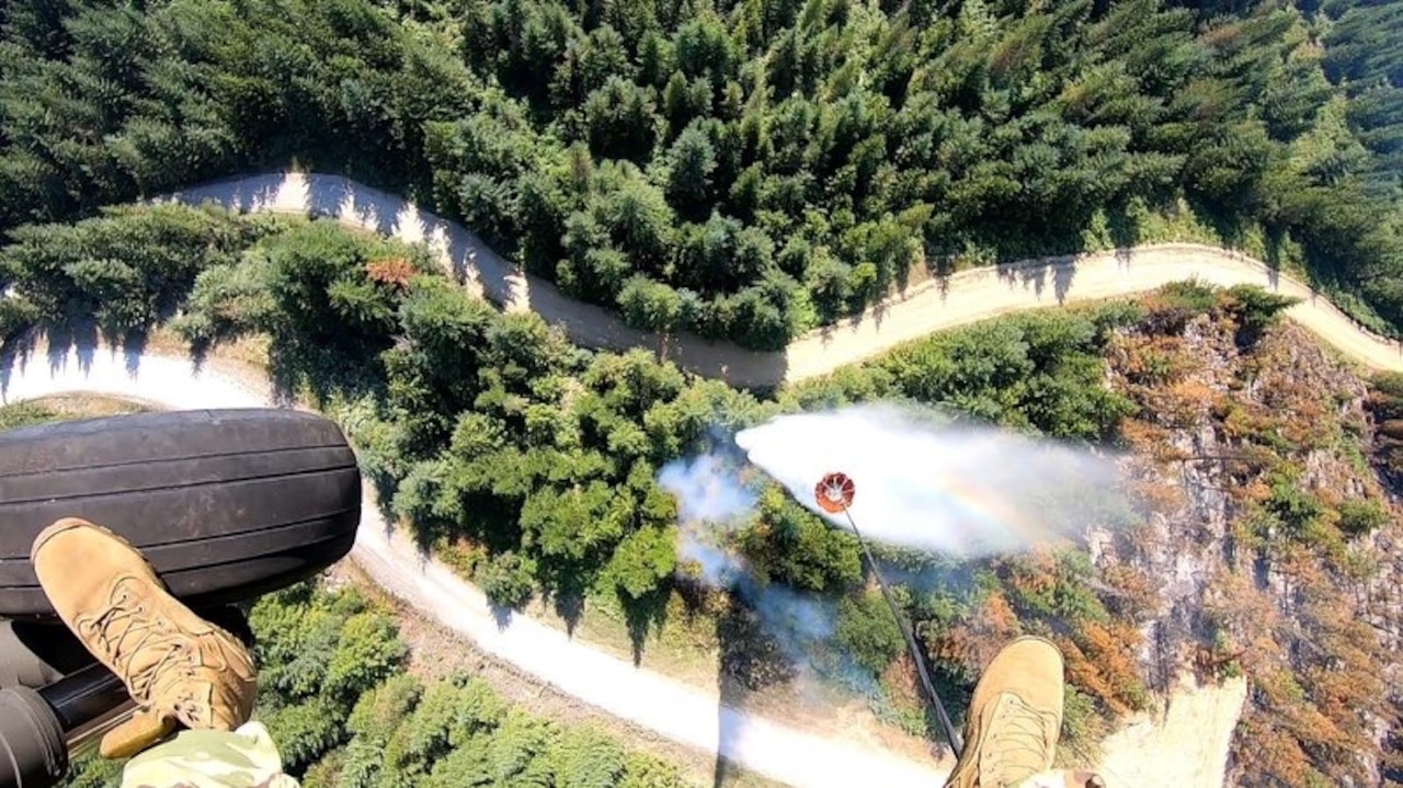 A person's legs dangle from a helicopter as it hovers over a smoky forest. A bucket is about to drop water on the ground.