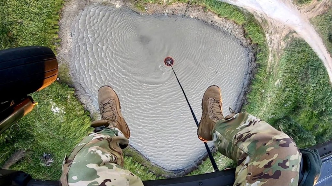 A person's legs dangle from a helicopter as it hovers above a lake where a bucket is dropped to collect water.