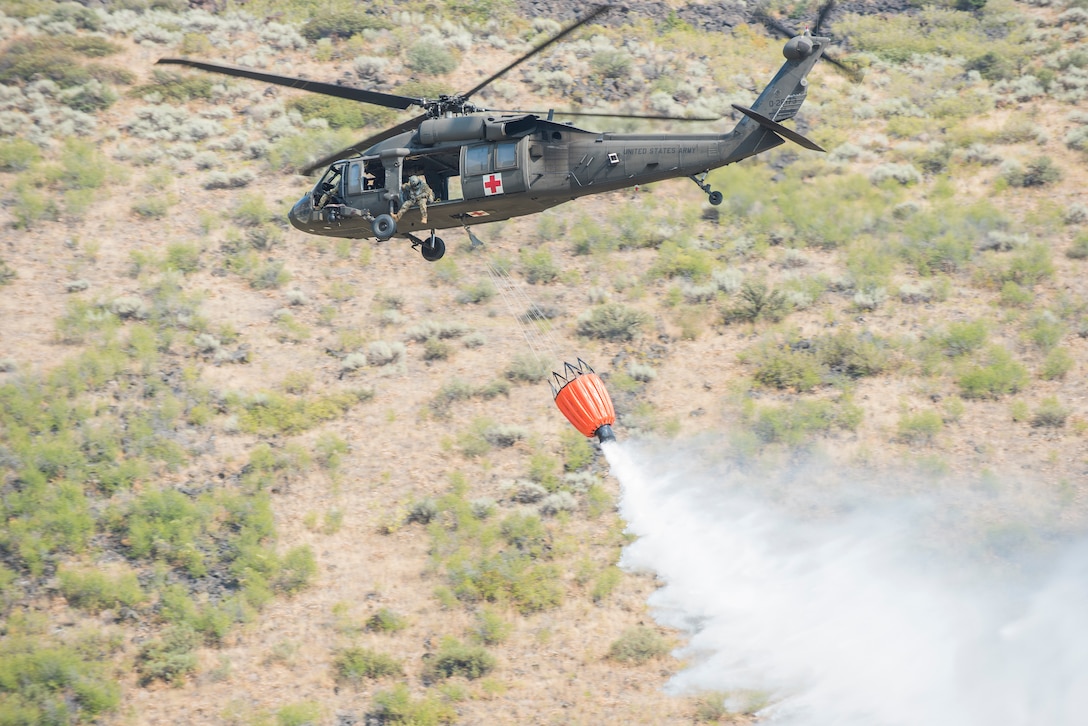 A large bucket attached to a helicopter drops water on the ground below.