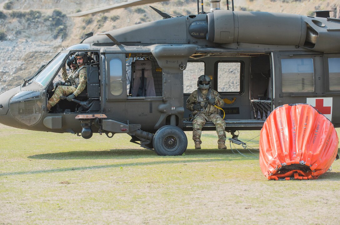 A helicopter and its crew sit grounded beside a large container with a small hole on top.