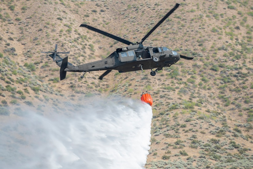 A large bucket attached to a helicopter drops water on the ground below.