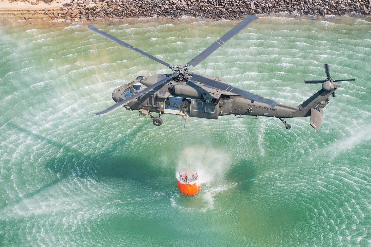 A helicopter lifts a large bucket out of a body of water.