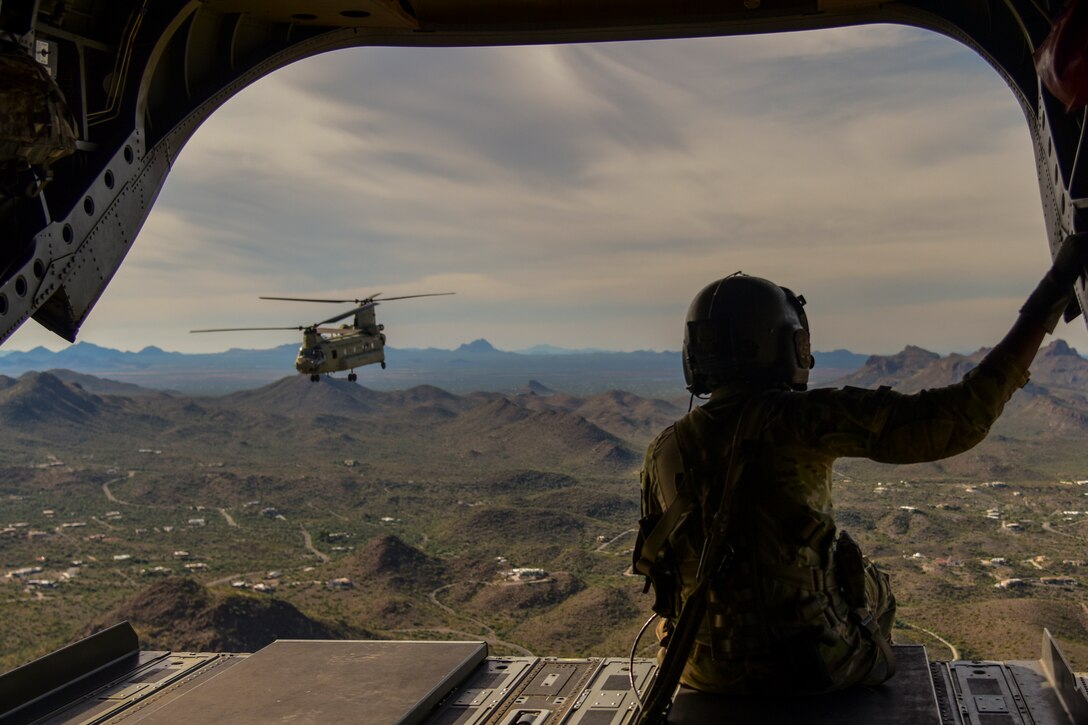 A soldier sits on the back of an airborne aircraft as another aircraft flies nearby with mountains in the background.
