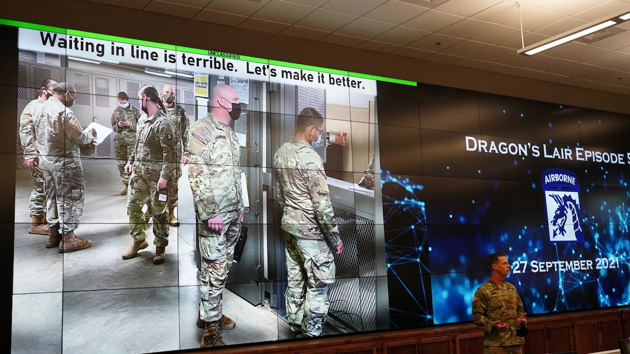 A man stands at the head of a room; images of soldiers are on a screen behind him.