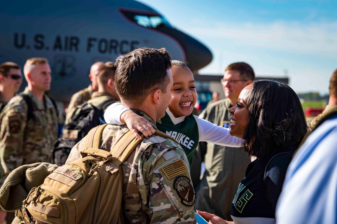 An airman hugs a child and a woman with other airmen and an aircraft in the background.