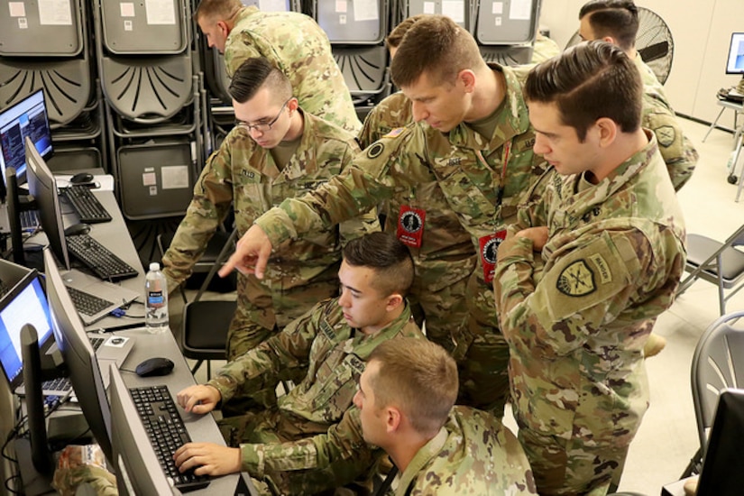 A group of national guardsmen huddle, some standing, around computer monitors.