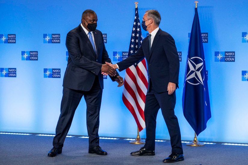 Secretary of Defense Lloyd J. Austin III shakes hands with another leader in front of NATO and U.S. flags.