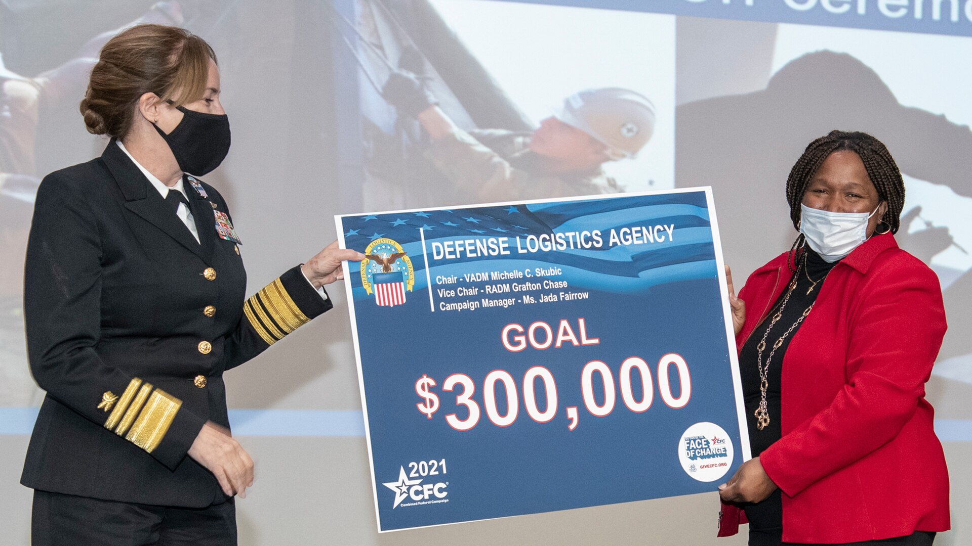 Two women - one in Navy dress uniform, one in a red jacket - stand holding a sign between them that says GOAL $300,000.