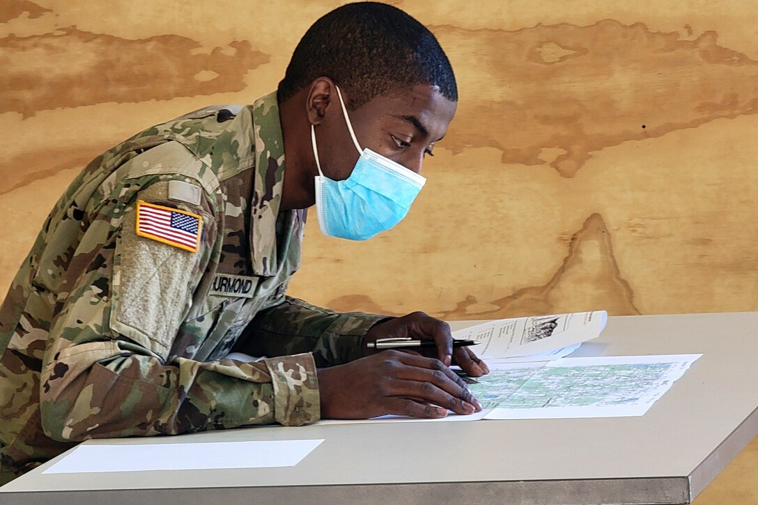A soldier wearing a mask sits at a table and looks at a map.