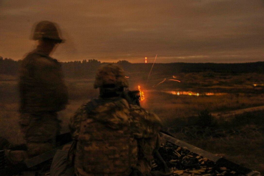 A soldier fires a weapon in a field at night as a fellow solider watches.