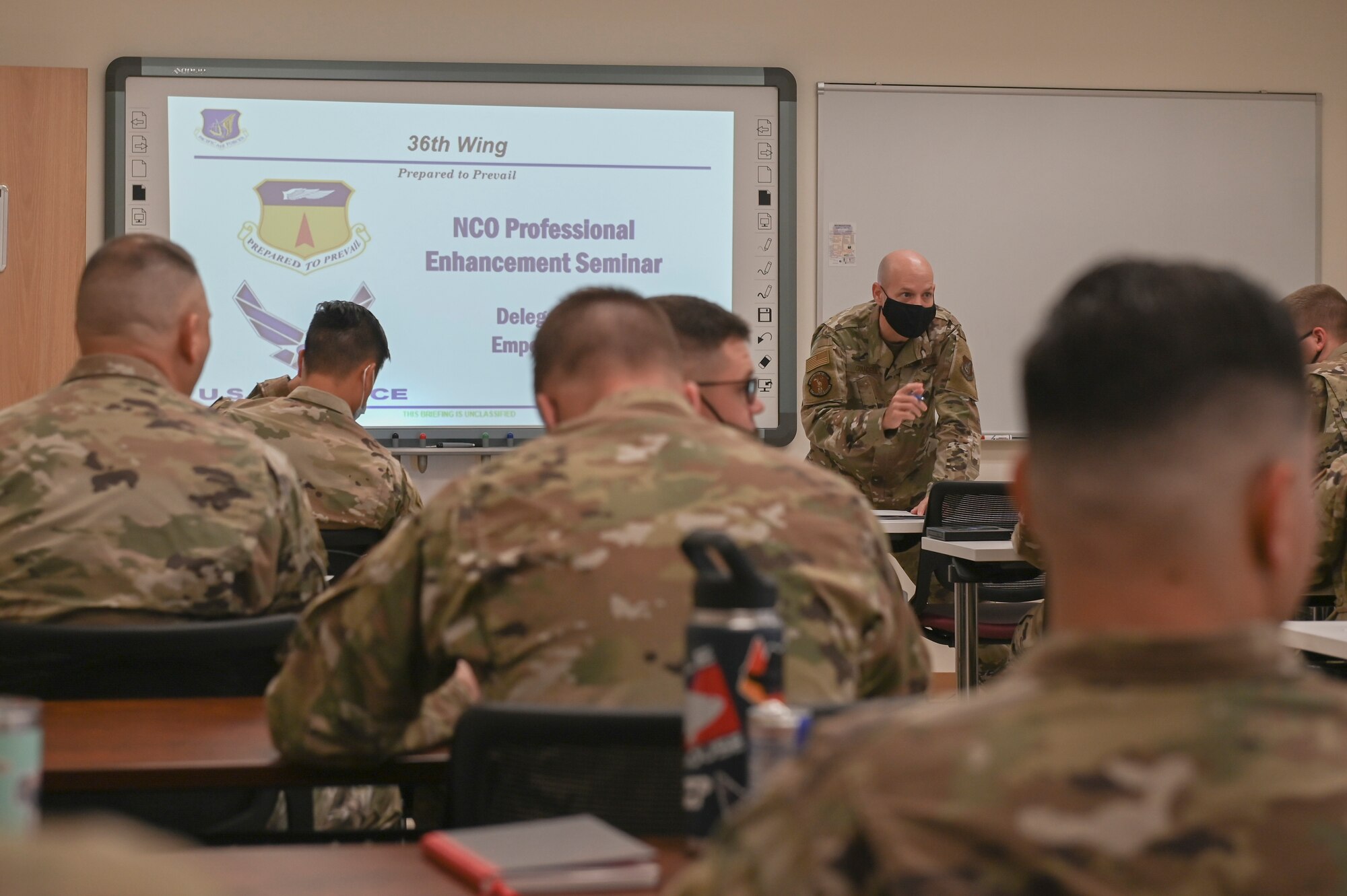A man stands at the front of a classroom, instructing service members.