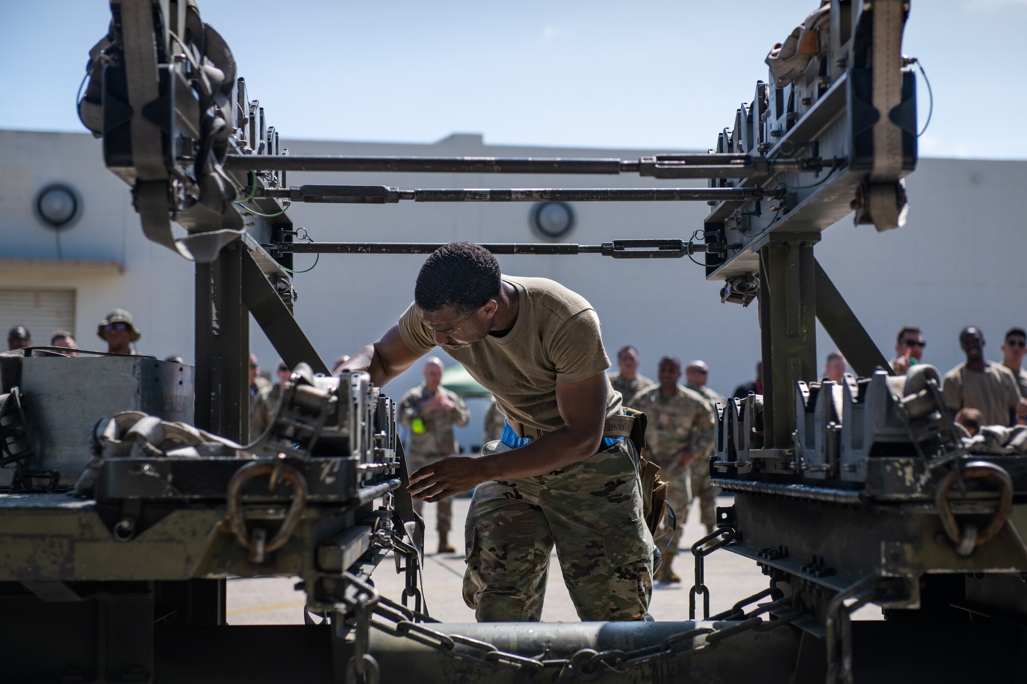 An airman stands framed by a munitions cart, securing straps on the cart
