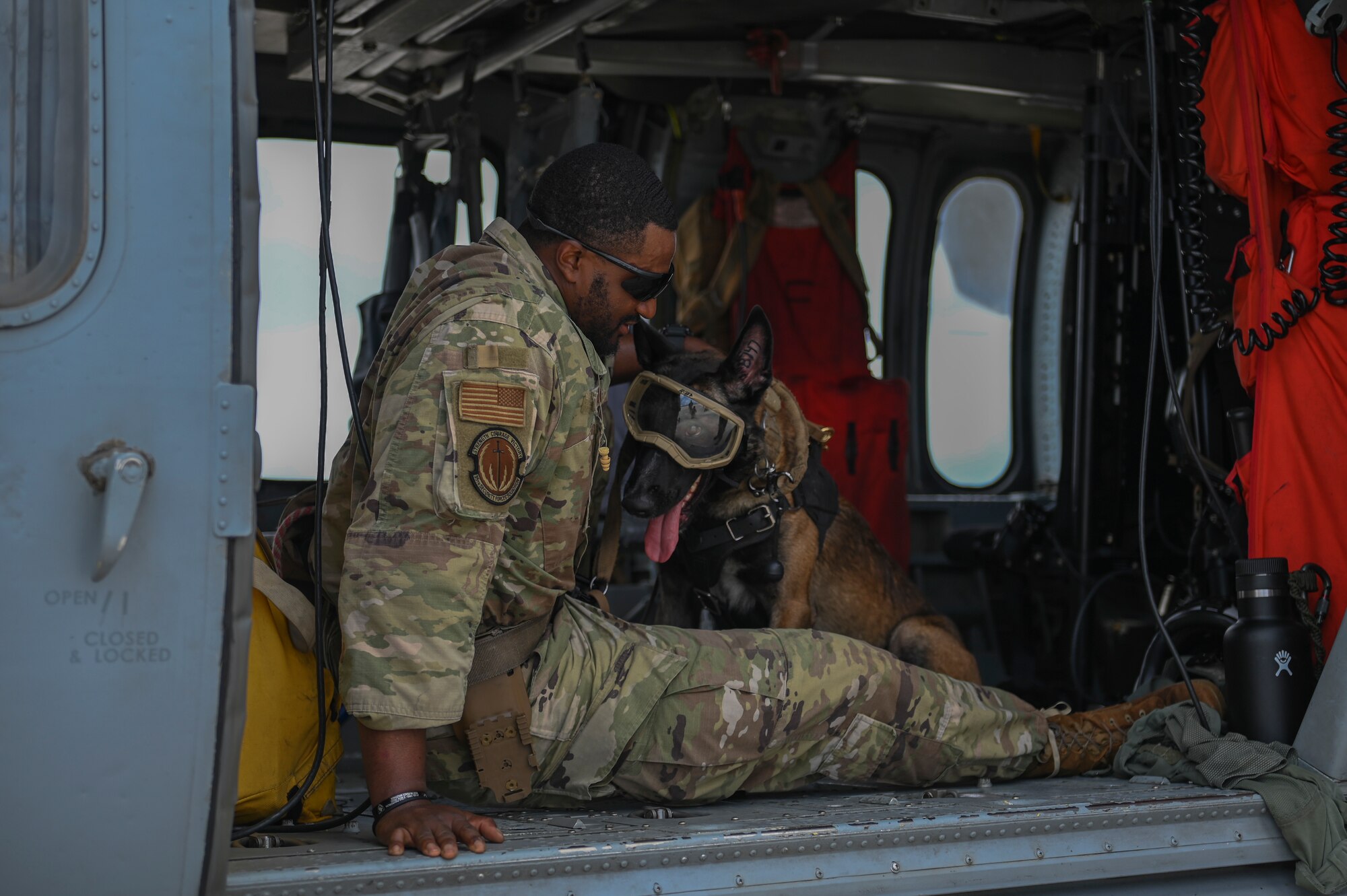 A military member sitting on the floor of a helicopter with a military working dog in front of him.