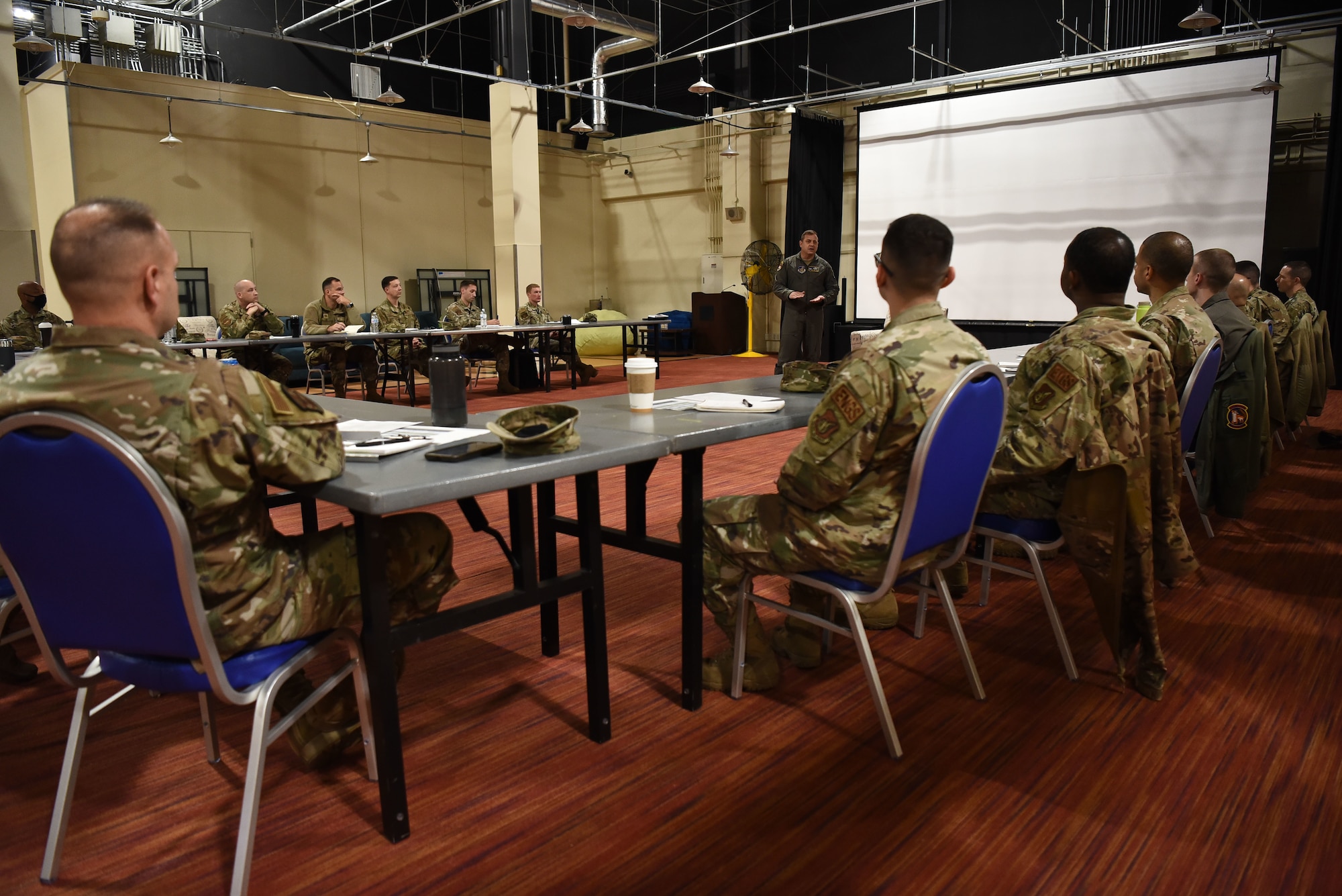 A military member in a jumpsuit talks to a class of military members.